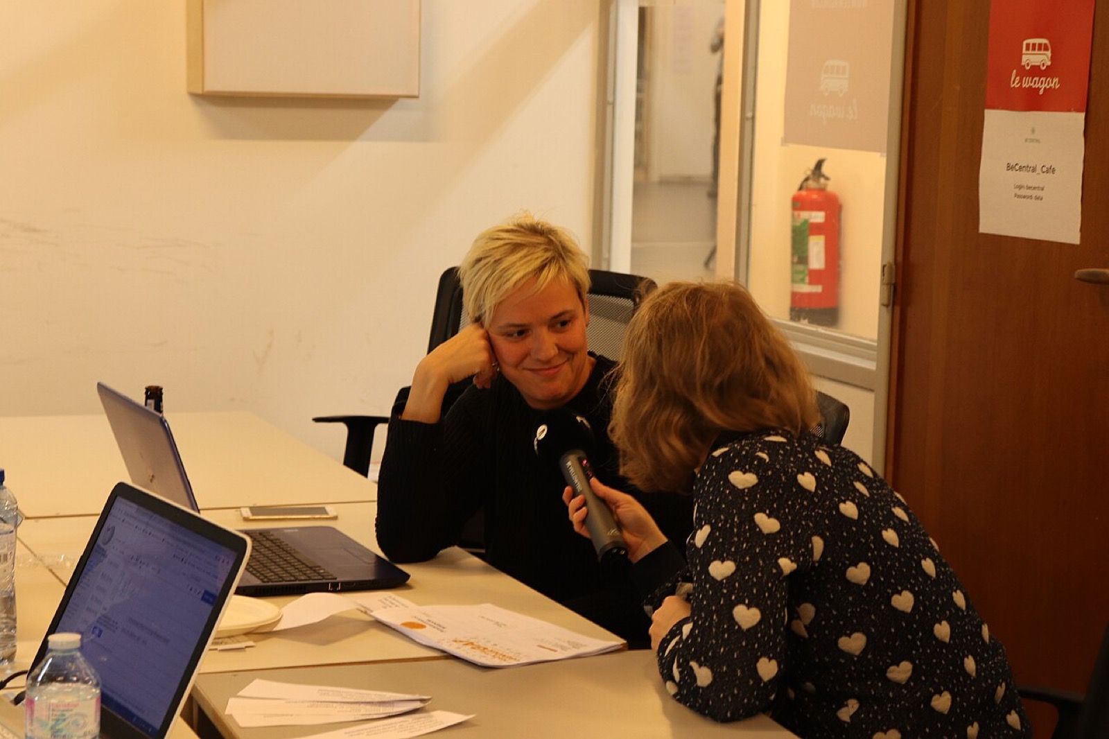 A real interview in which an interviewer holds a microphone while speaking with a participant, with a computer and notes on the table